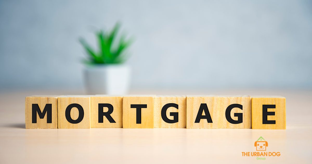 Wooden letter blocks spelling “Mortgage” on a table with a blurred plant in the background and The Urban Dog Group logo in the corner.